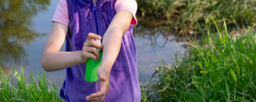 Ein Kind, welches sich den Arm mit Mückenschutzspra einsprüht. IM Hintergrund sieht man viel grün, das Kind ist in der Natur.